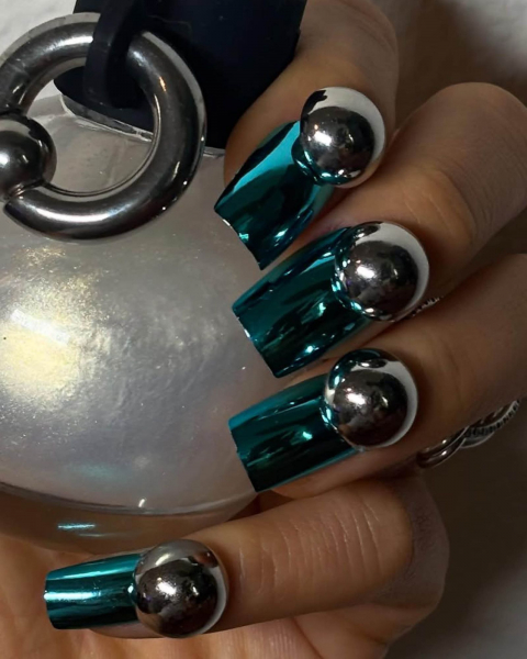 Turquoise nails with large silver balls at the base clutching a pearly nail polish bottle.